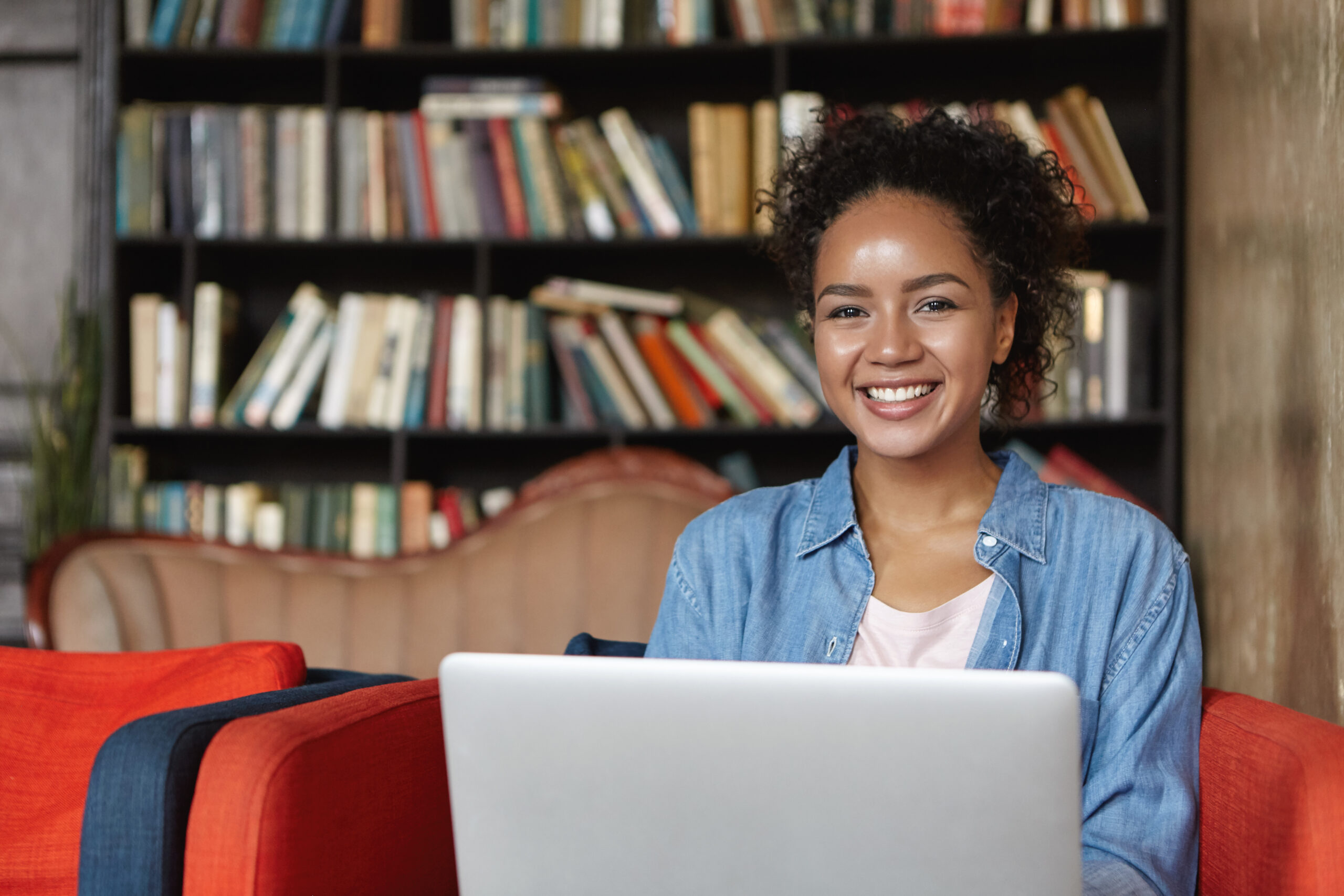 Ragazza allegra e sorridente è seduta su un divano appoggiato a scaffali con libri mentre lavora utilizzando un computer portatile.